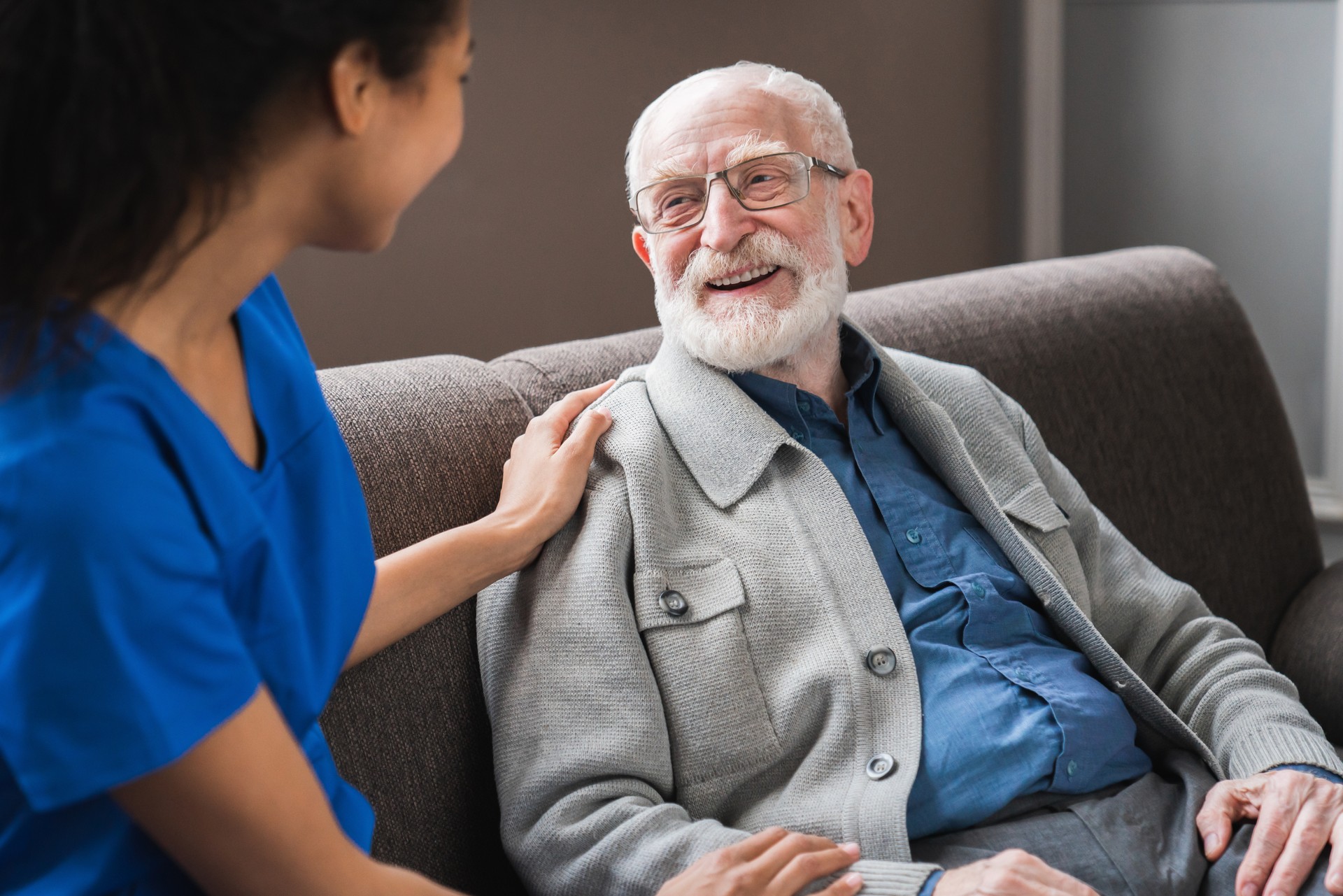 Young friendly caregiver talking chatting to happy senior man in hallway of nursing home. Picture of smiling nurse assisting senior elderly man